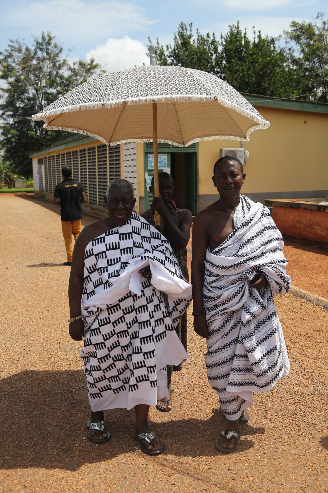 Traditional leaders in woven cloth walking beneath a ceremonial umbrella